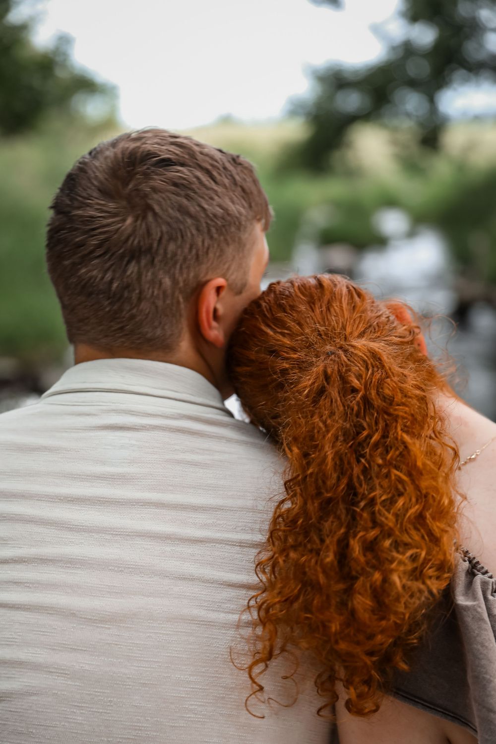 A woman with curly red hair rests her head on a man's shoulder outdoors near a river.