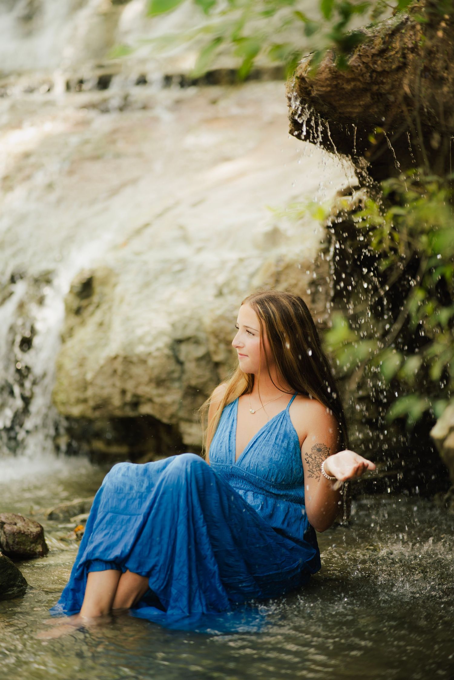 Young woman in blue dress sitting in shallow water near a waterfall surrounded by lush greenery.