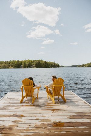 Couple sitting in yellow Adirondack chairs on a wooden dock overlooking a serene lake surrounded by trees.