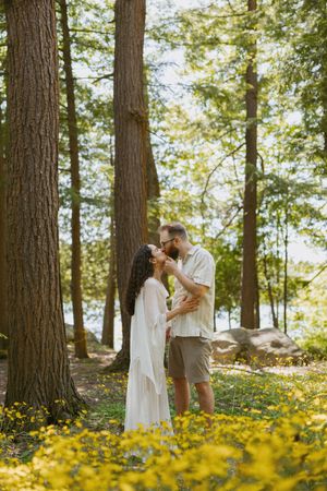 Couple sharing a romantic kiss in a sunlit forest clearing with yellow wildflowers and tall pine trees.