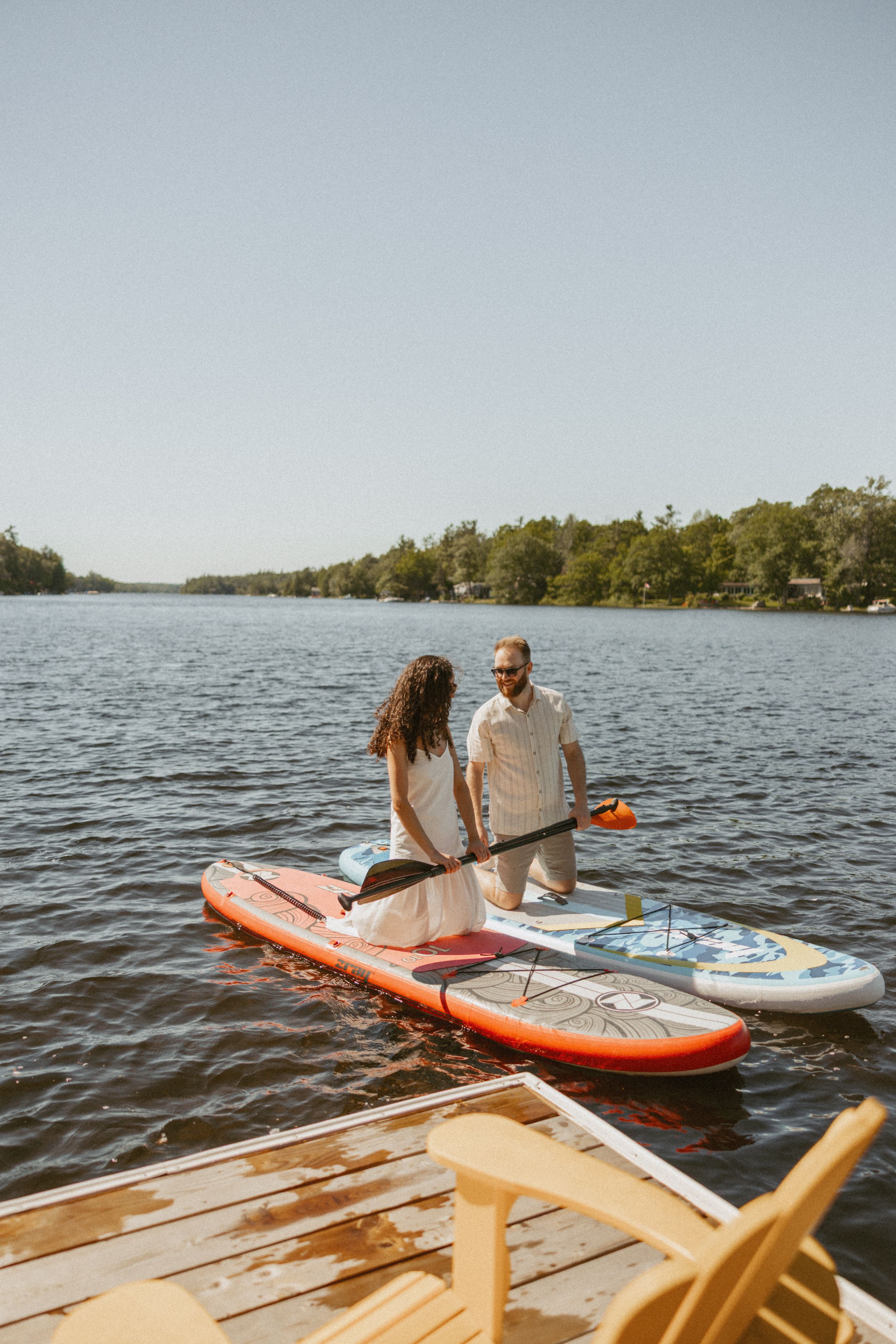 Couple standing on a dock with paddleboards on a calm lake surrounded by trees on a sunny day.