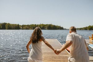 Couple holding hands on a sunny wooden dock with a calm lake and lush green trees in the background.