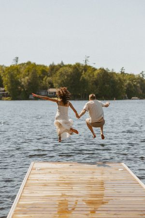 Couple holding hands jumping off a wooden dock into a lake surrounded by lush green trees on a sunny day.