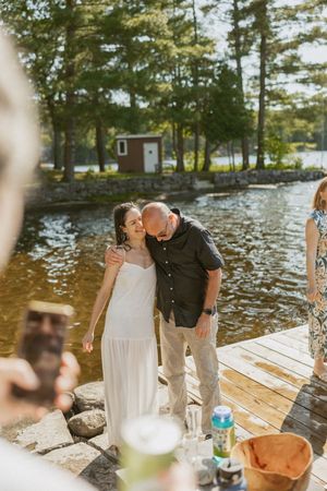 Couple sharing a kiss on a lakeside dock in summer, woman in white dress, man in black shirt, trees reflected in water.