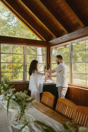 Couple in white attire toasting in a sunlit wood-paneled cabin dining room surrounded by lush green trees.