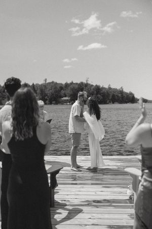 Black and white photo of a couple kissing on a lakeside dock during an outdoor wedding ceremony with guests watching.
