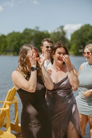 Two women in elegant dresses laugh and cry tears of joy outdoors near a lake at a sunny summer celebration.
