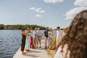 Group of people gathered on a wooden dock by a lake on a sunny day, surrounded by lush green trees.