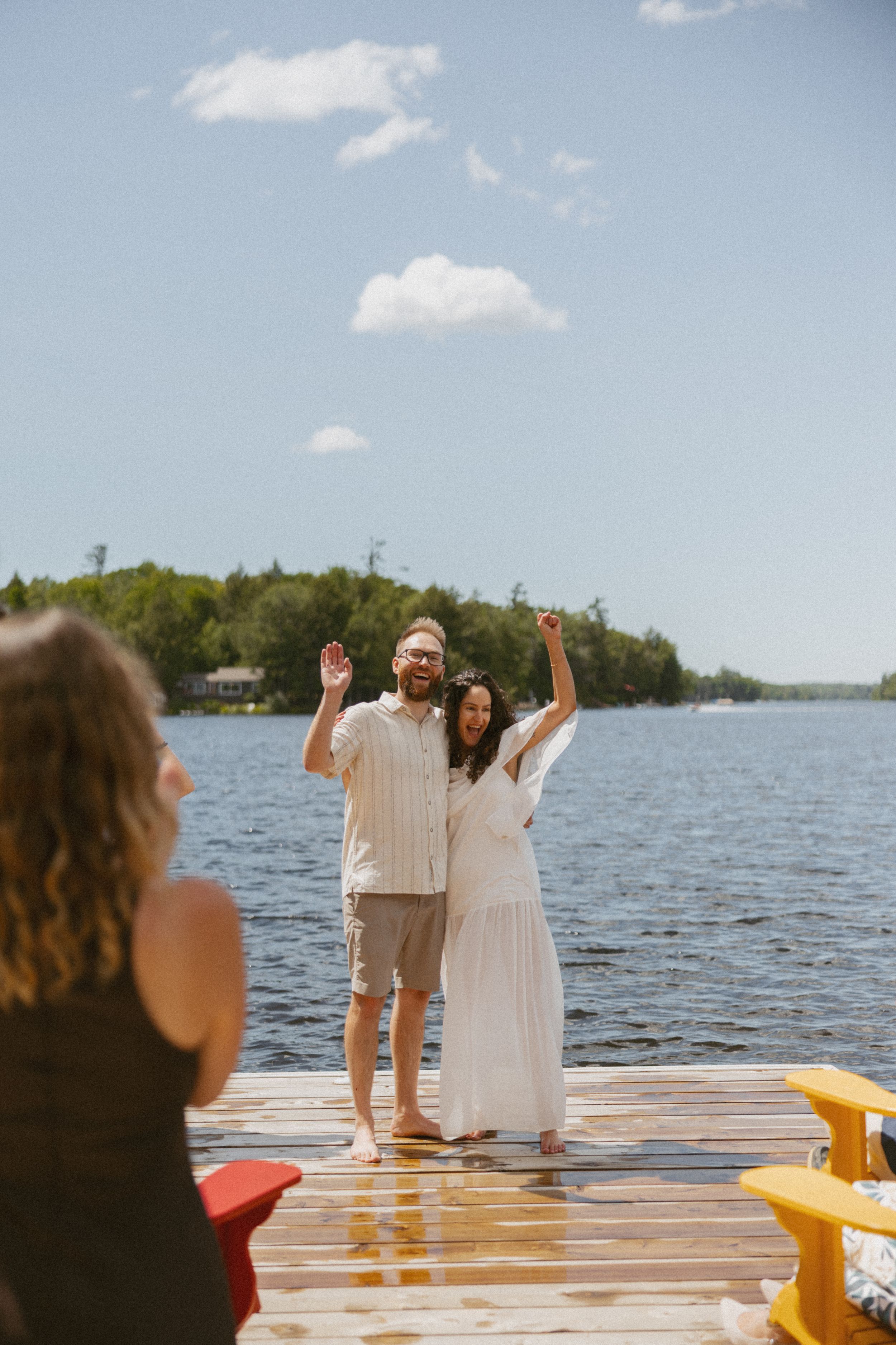 Happy couple celebrating on a sunny lakeside dock, woman in white dress and man in casual attire, raising hands joyfully.