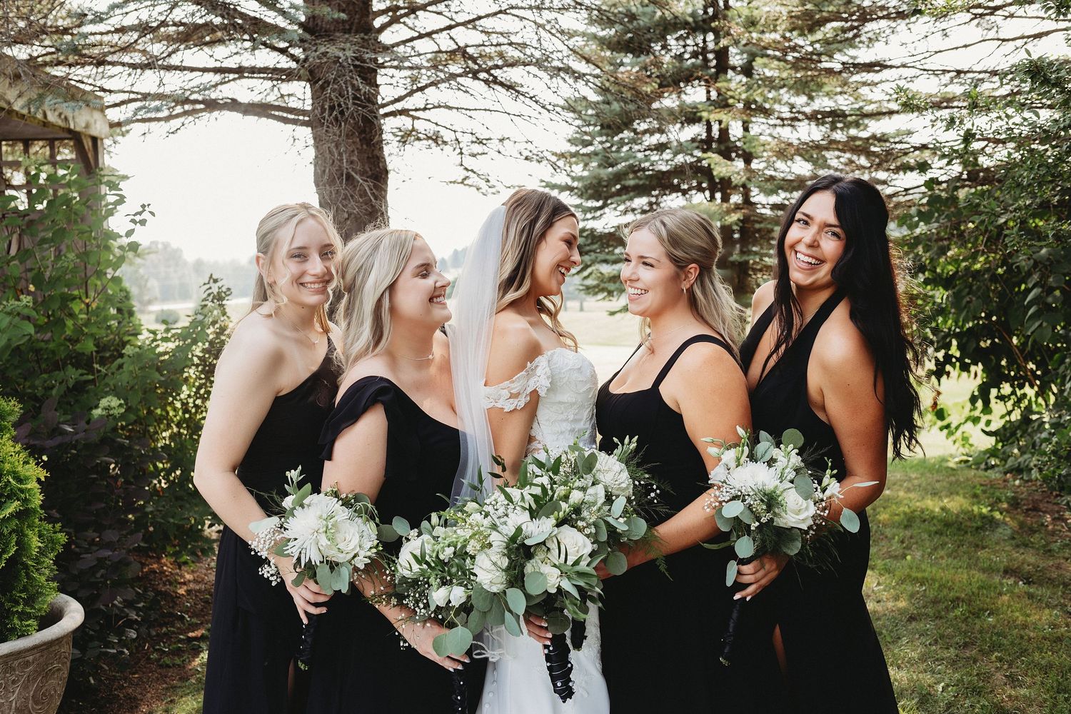 Bride laughing with four bridesmaids wearing black dresses and holding white and green bouquets outdoors.