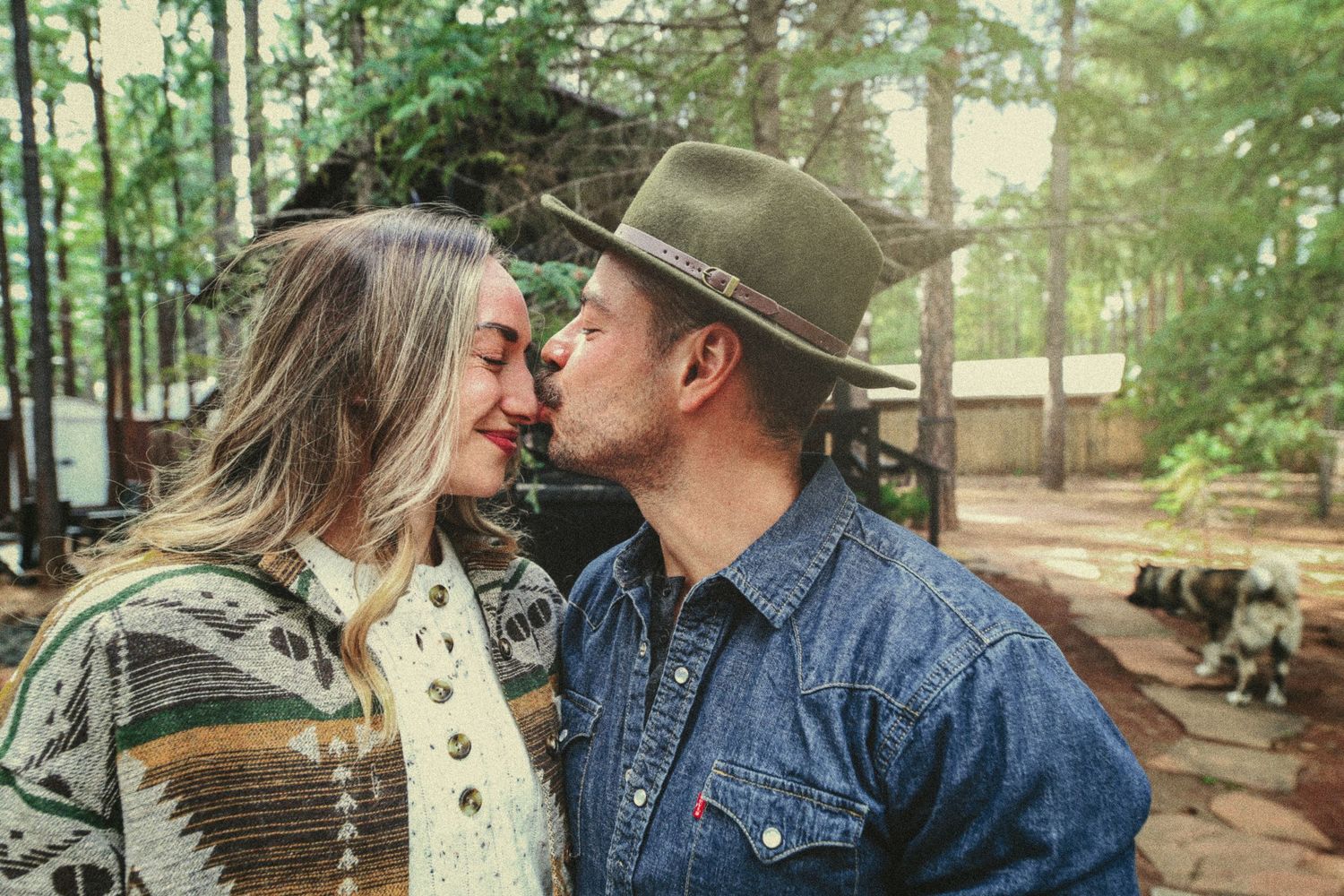 A couple shares a tender nose-to-nose moment outdoors among tall trees, he in a green hat and denim shirt, she in a patterned cardigan.