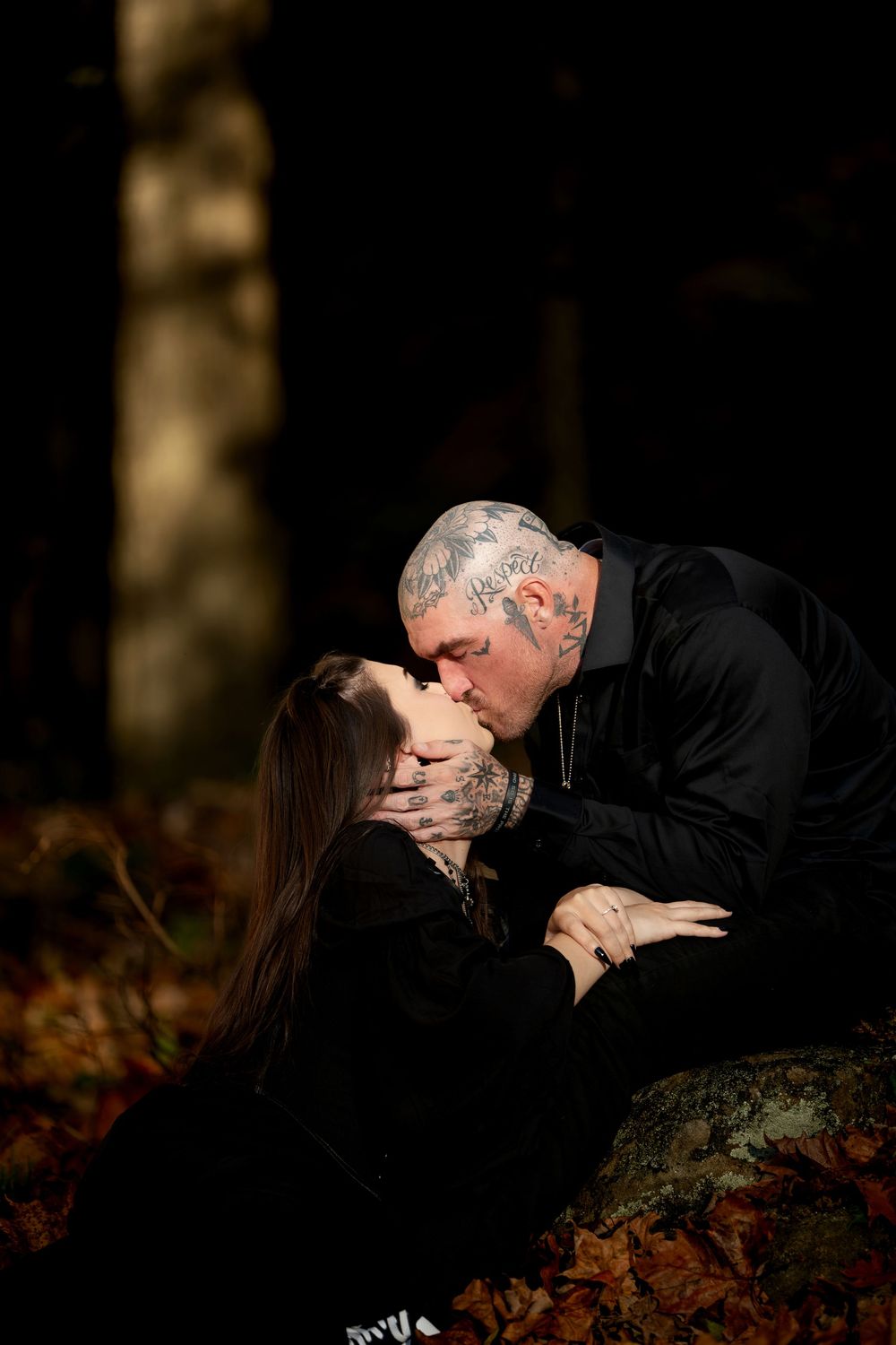 Tattooed couple sharing a romantic kiss on a mossy rock in a dark autumn forest with fallen leaves.