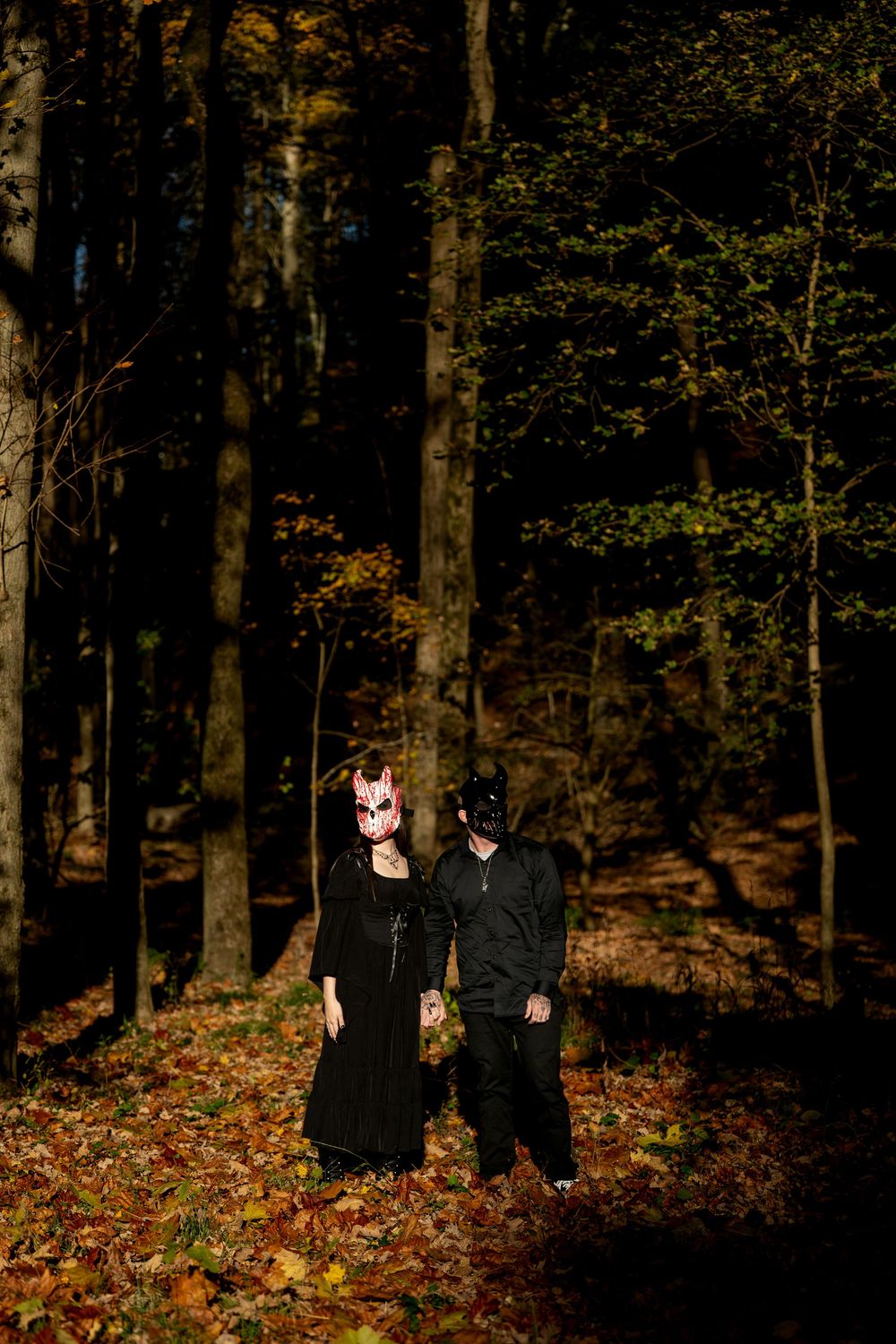 Two people in dark clothing and animal masks stand among fallen autumn leaves in a shadowy forest lit by golden sunlight.
