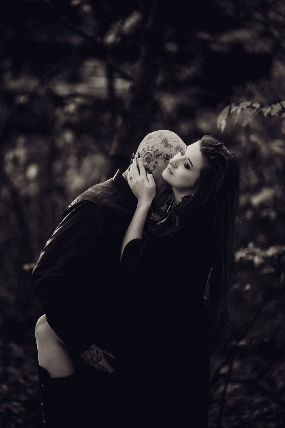 Black and white portrait of a woman embracing a tattooed bald man in a dark, moody forest setting.