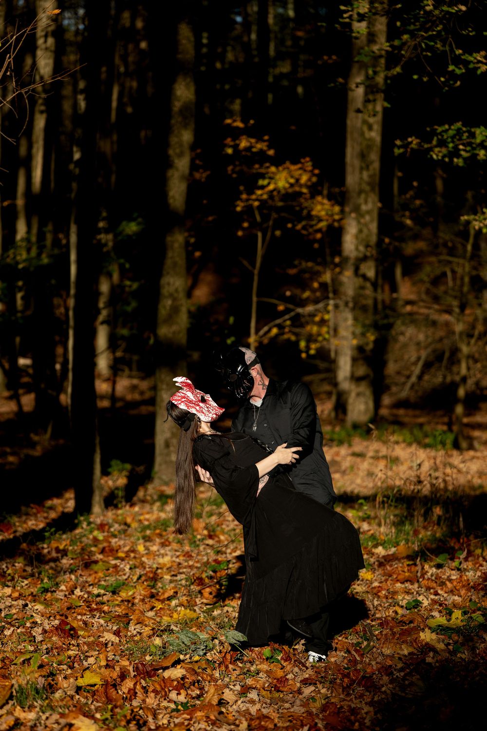 A couple in black embrace in a dramatic dip in an autumn forest, the woman wearing a red and white mask.