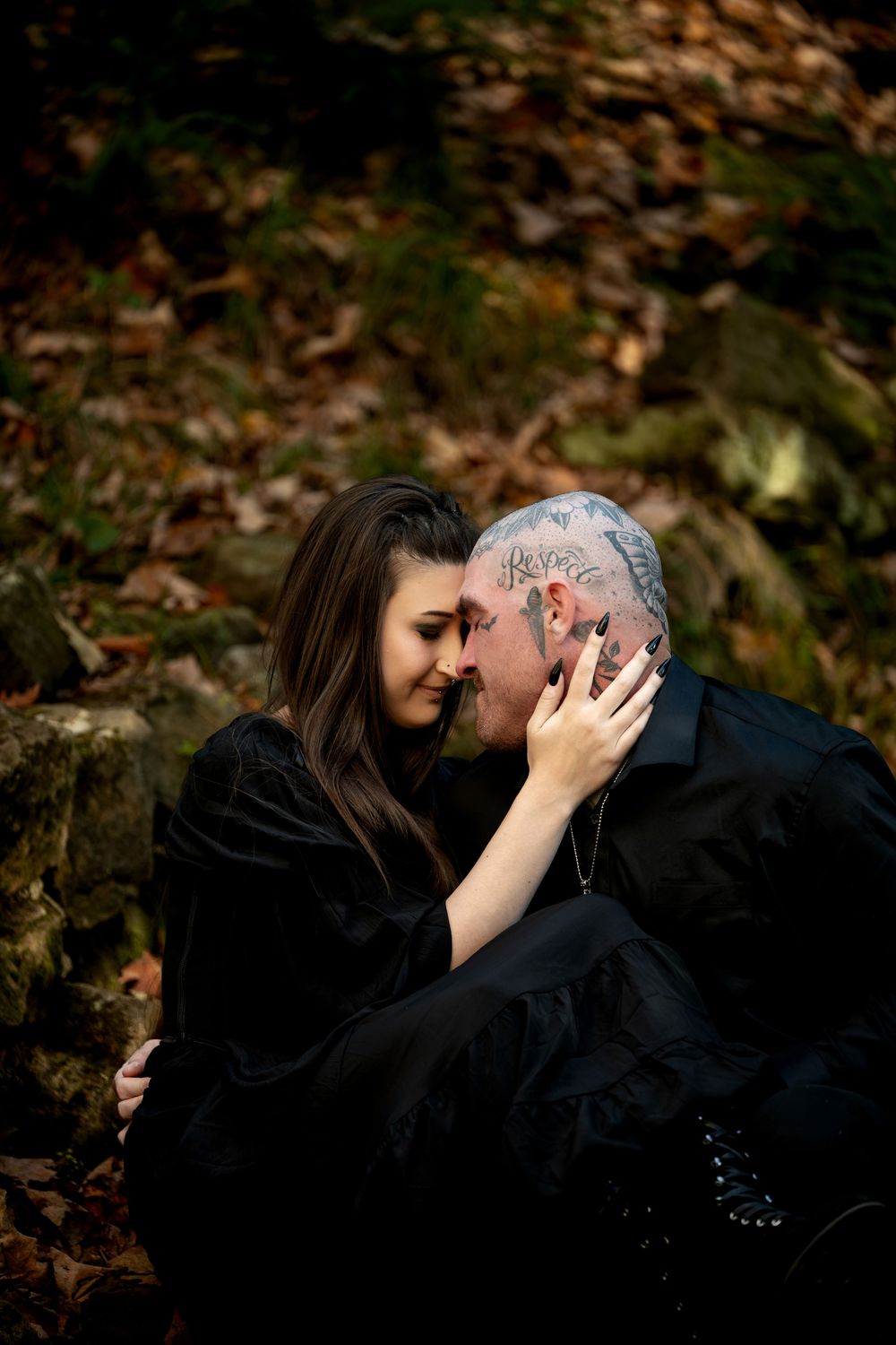 Woman in black gently touching foreheads with a tattooed man in a romantic embrace on a mossy, leaf-covered forest hillside.