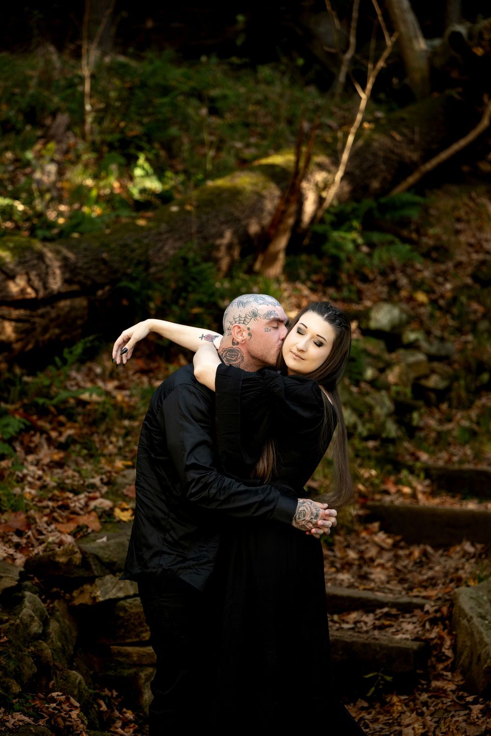 Tattooed man in black kisses the forehead of a woman in a black dress as they embrace on a mossy forest path with fallen autumn leaves.