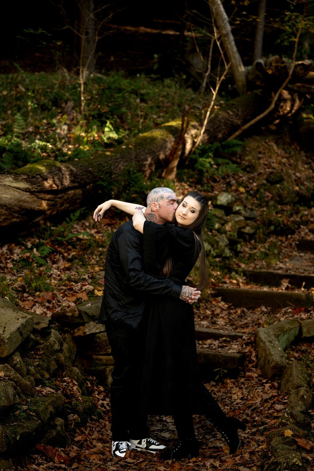 Tattooed man kisses the cheek of a woman in a black dress as they embrace on mossy stone steps in an autumn forest.