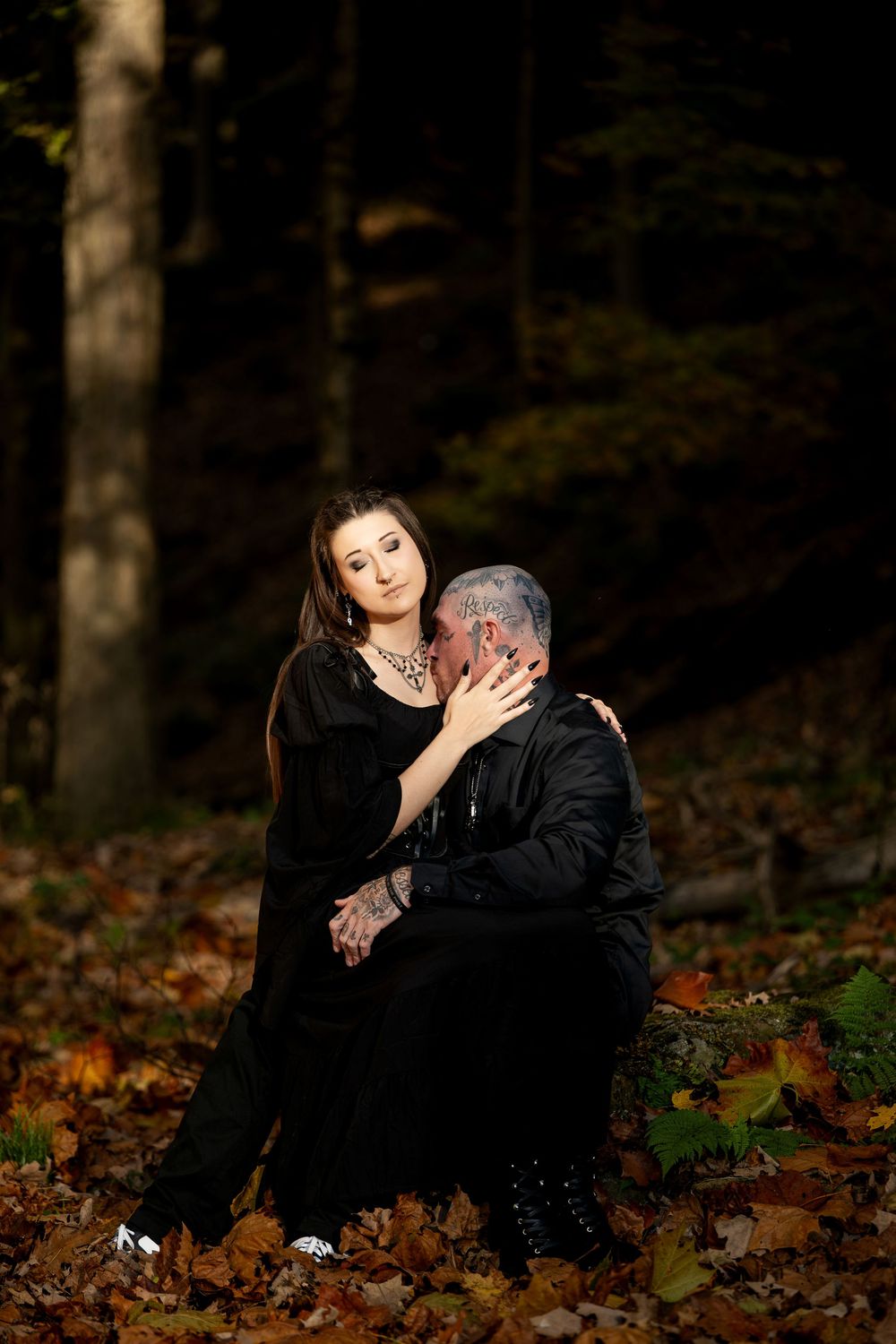 A couple in dark clothing embrace while seated on a log in an autumn forest, surrounded by fallen orange leaves.