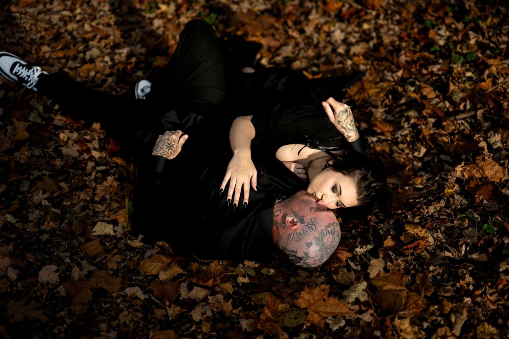 A couple in black lie close together on a bed of fallen autumn leaves, the woman gazing at the camera in a moody engagement photo.