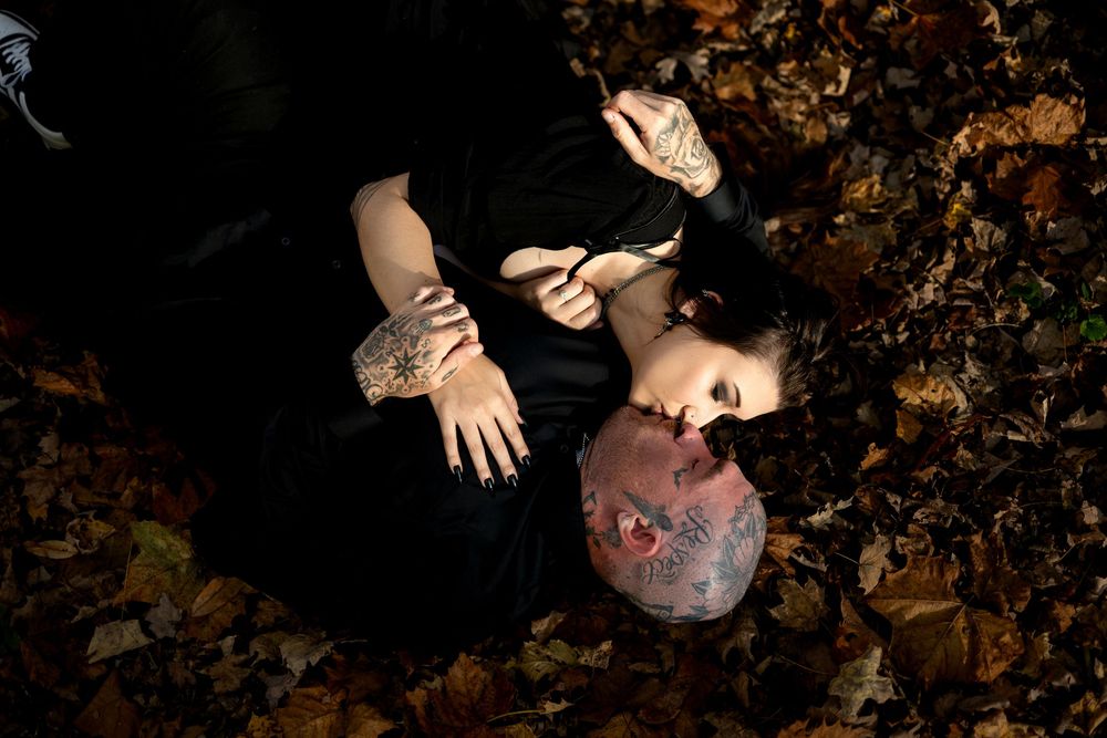 A tattooed man and a woman in black lie entwined on autumn leaves, their hands clasped in a dramatic overhead couple portrait.