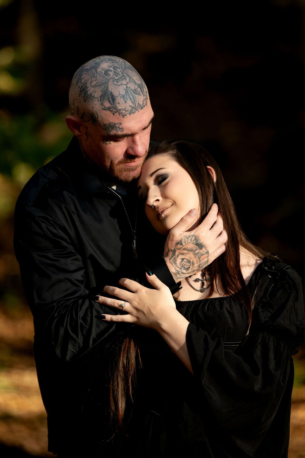 Woman in a black gown rests her head on a tattooed man's chest as they embrace in a golden-lit autumn forest.