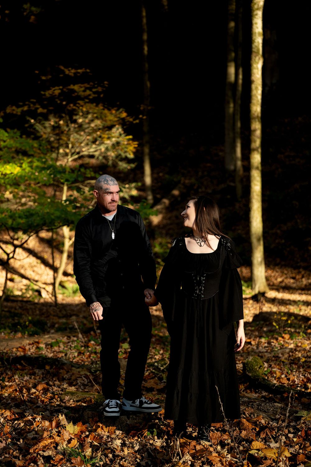 A couple in black gothic attire stand together in a sunlit autumn forest, smiling at each other among fallen leaves.