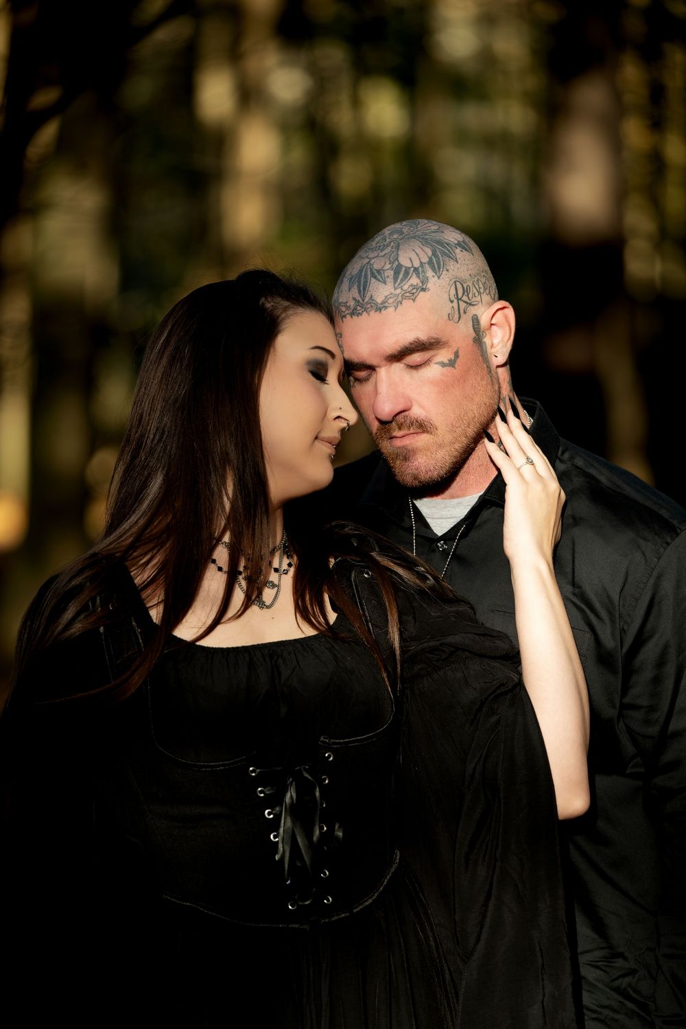 Close portrait of a woman in a black corset dress nuzzling a man with a tattooed head in moody forest light.