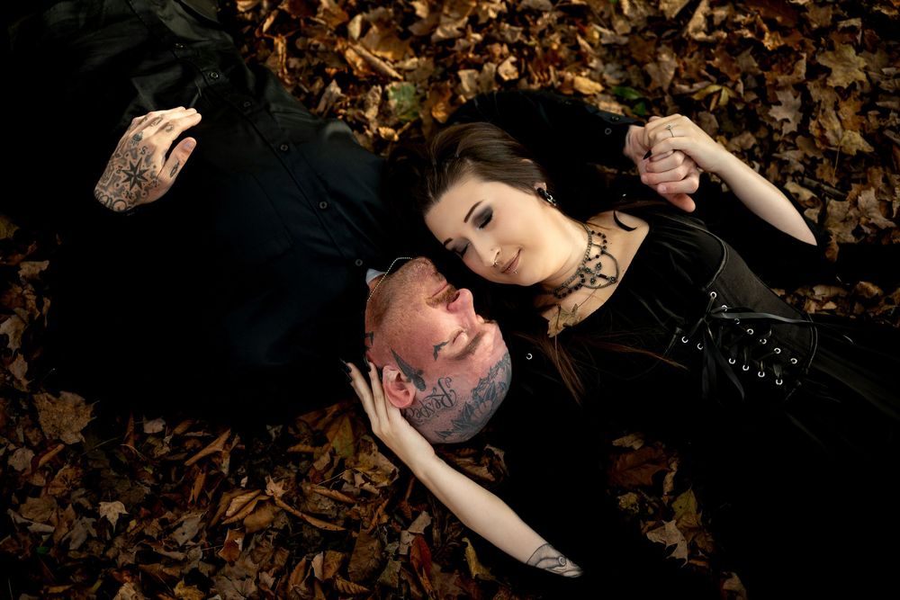Overhead view of a couple in black lying together on fallen autumn leaves, his tattoos visible as they gaze at each other.