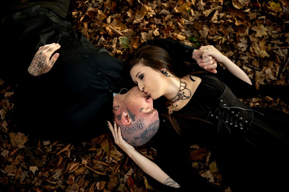 A woman in a black gothic outfit kisses the forehead of a tattooed man lying among colorful autumn leaves.