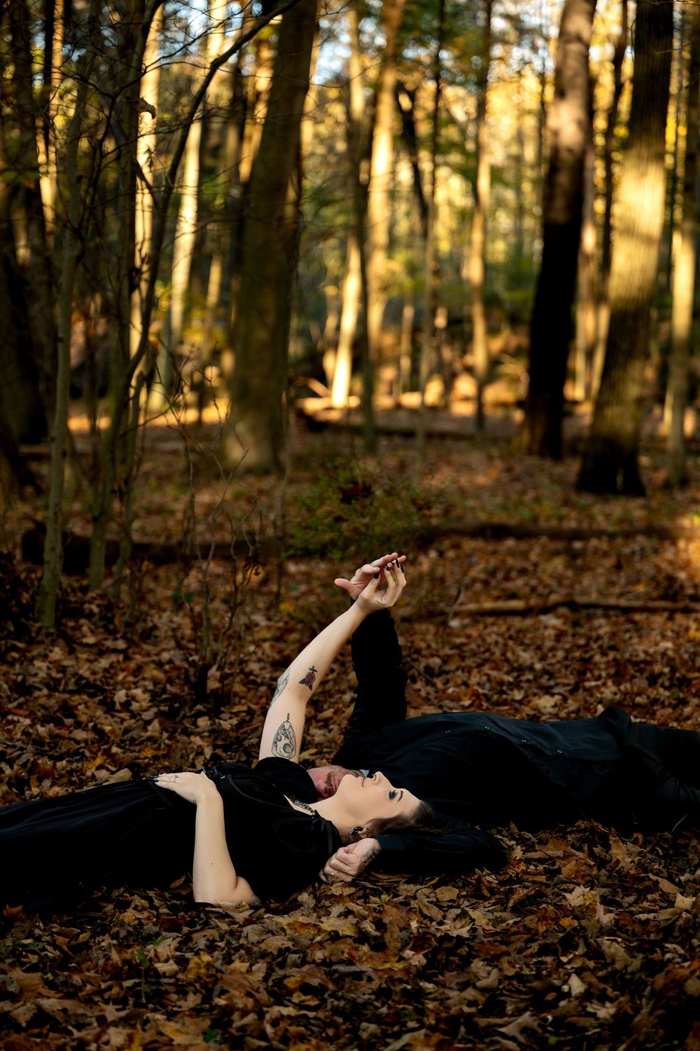 Two people in black clothing lie on a bed of fallen autumn leaves in the woods, one reaching an arm toward the sky.
