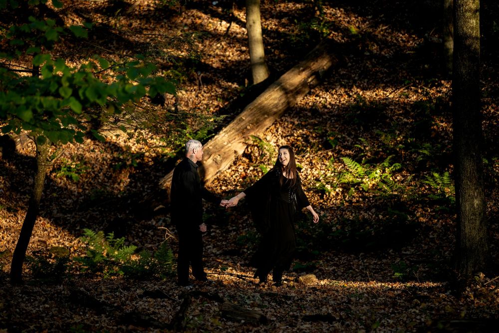 A couple in black clasp hands as they walk among tall trees and fallen leaves in a dark, sun-dappled autumn forest.