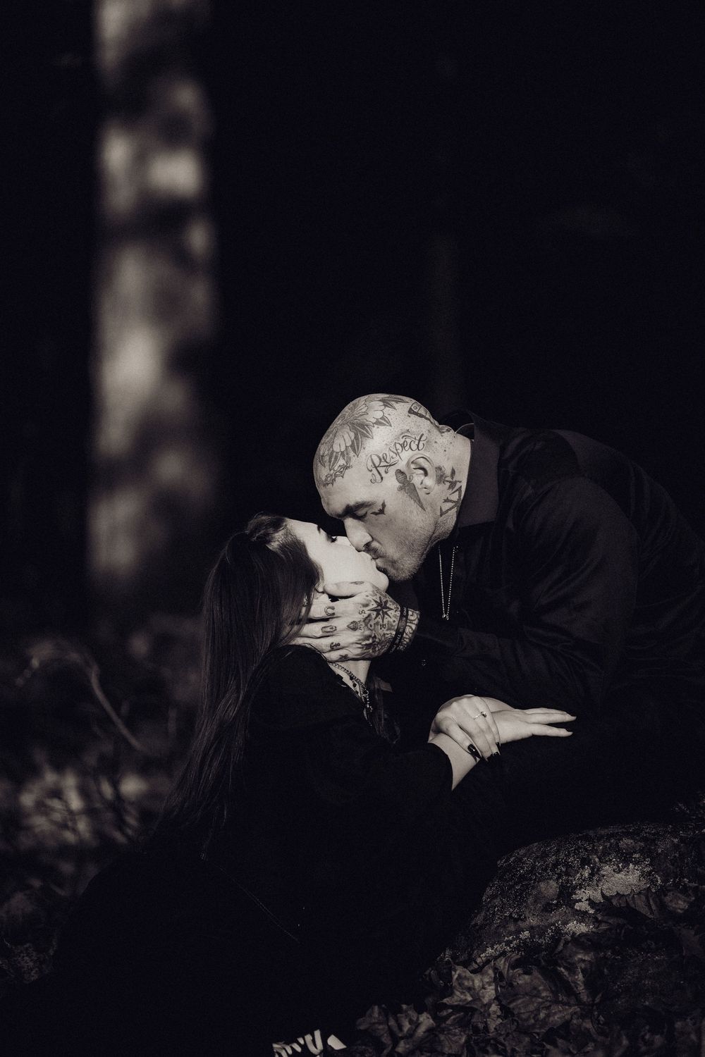 Black and white portrait of a tattooed man kissing a woman in dark clothing, leaning over her in a moody forest setting.