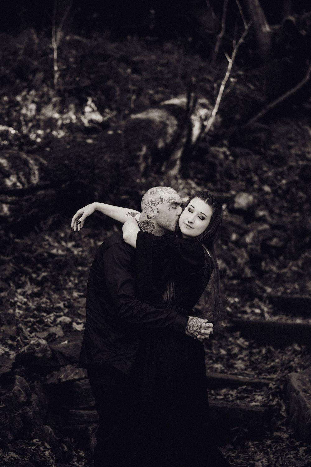 A black and white image of a tattooed man embracing a woman in dark clothing on a leaf-covered forest path.