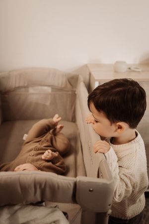 Young toddler boy leaning over a baby bassinet looking at a newborn sibling dressed in brown.