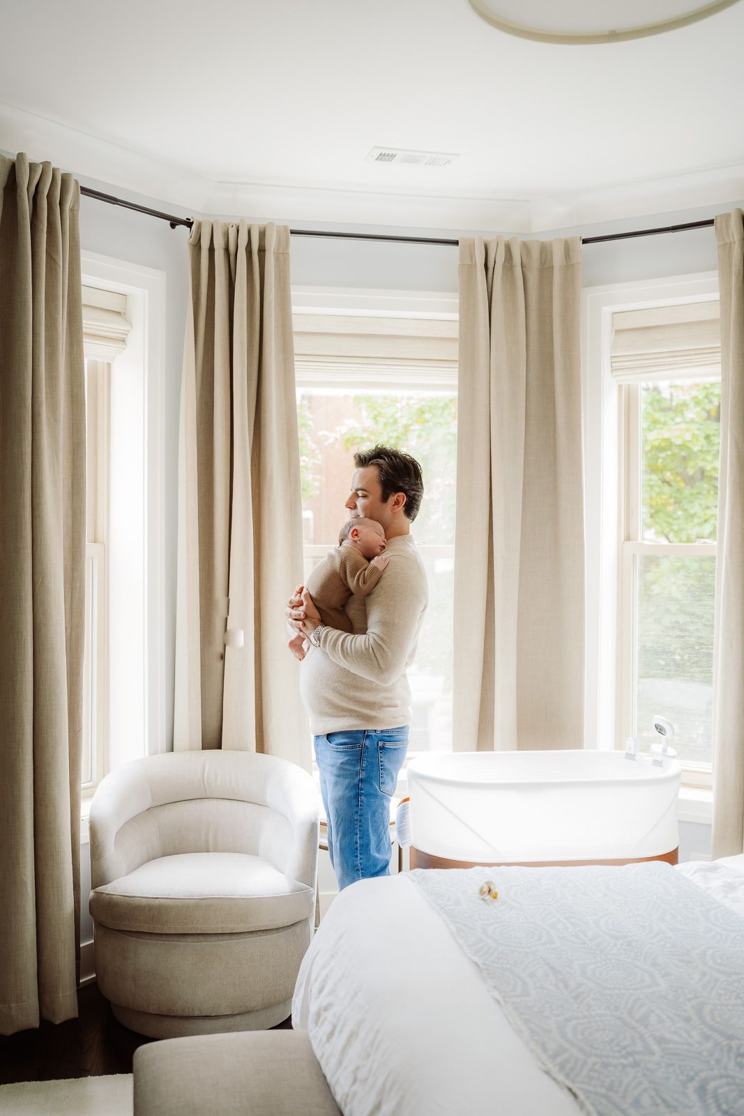 A parent holds a newborn baby while standing by bay windows in a softly decorated nursery bedroom.