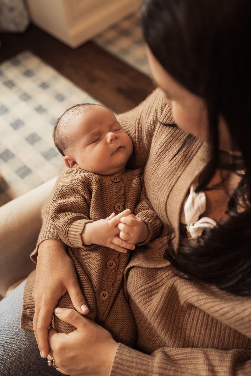 A mother in a brown knit cardigan tenderly holds her sleeping newborn baby dressed in a matching brown button-up outfit.