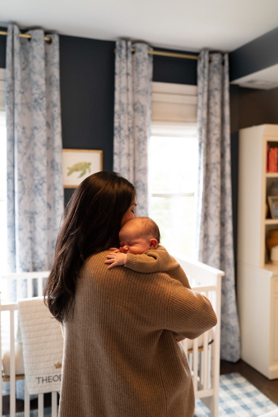 Mother holding newborn baby in a navy blue nursery with floral curtains and white crib.