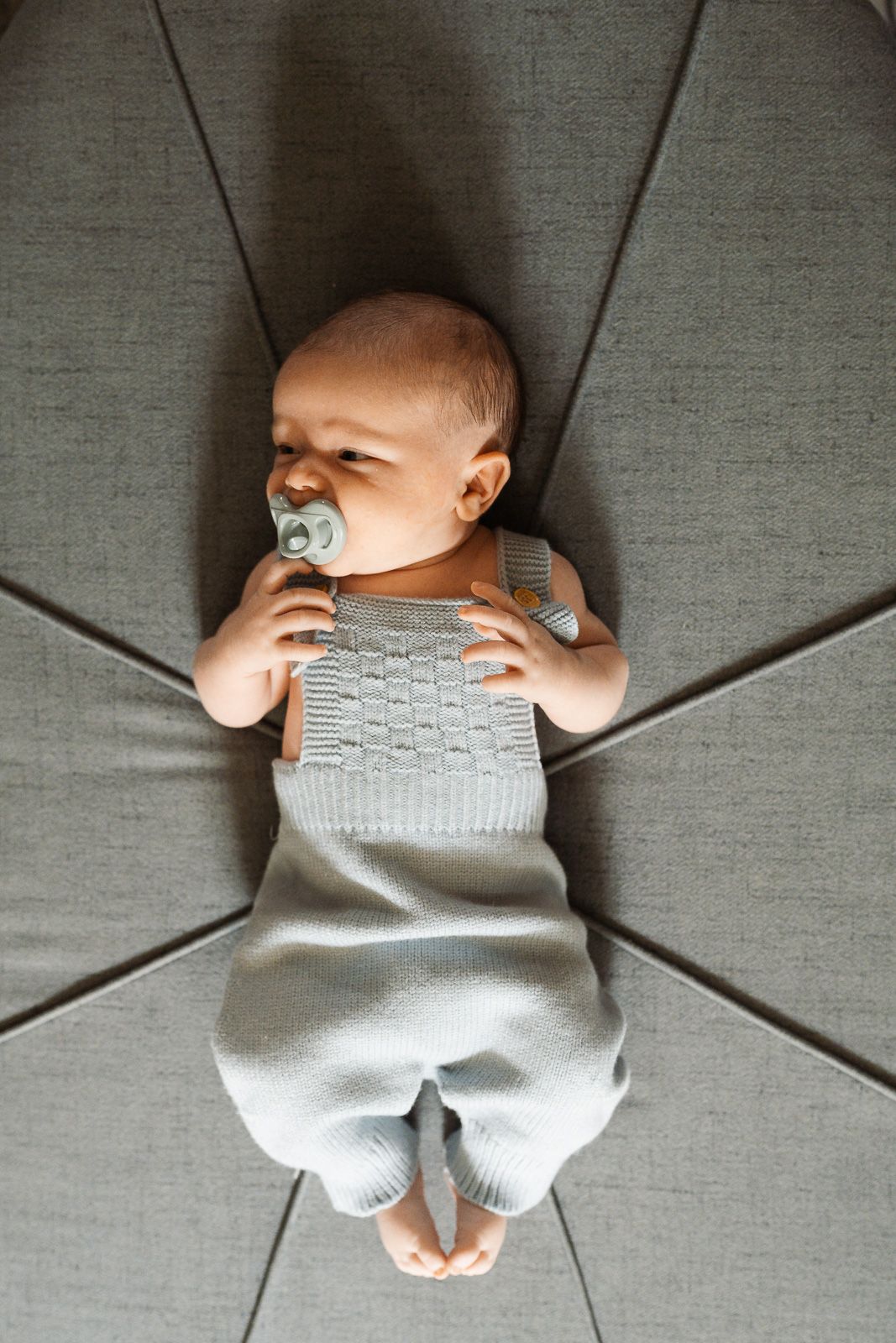 Adorable baby in gray knit overalls lying on a cushioned chair, holding a pacifier with bare feet visible.
