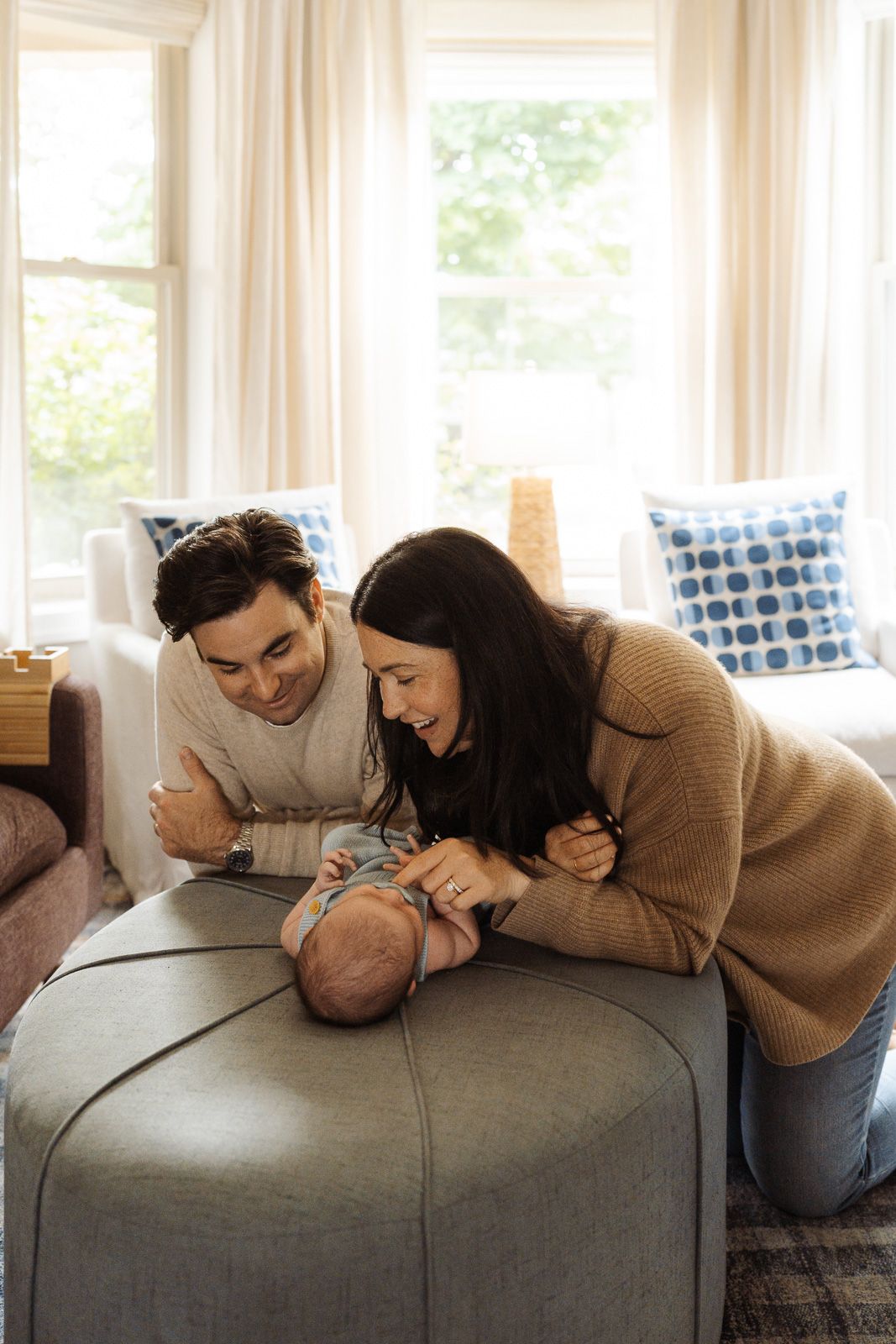 Happy parents lean over an ottoman admiring their newborn baby in a bright, cozy living room.