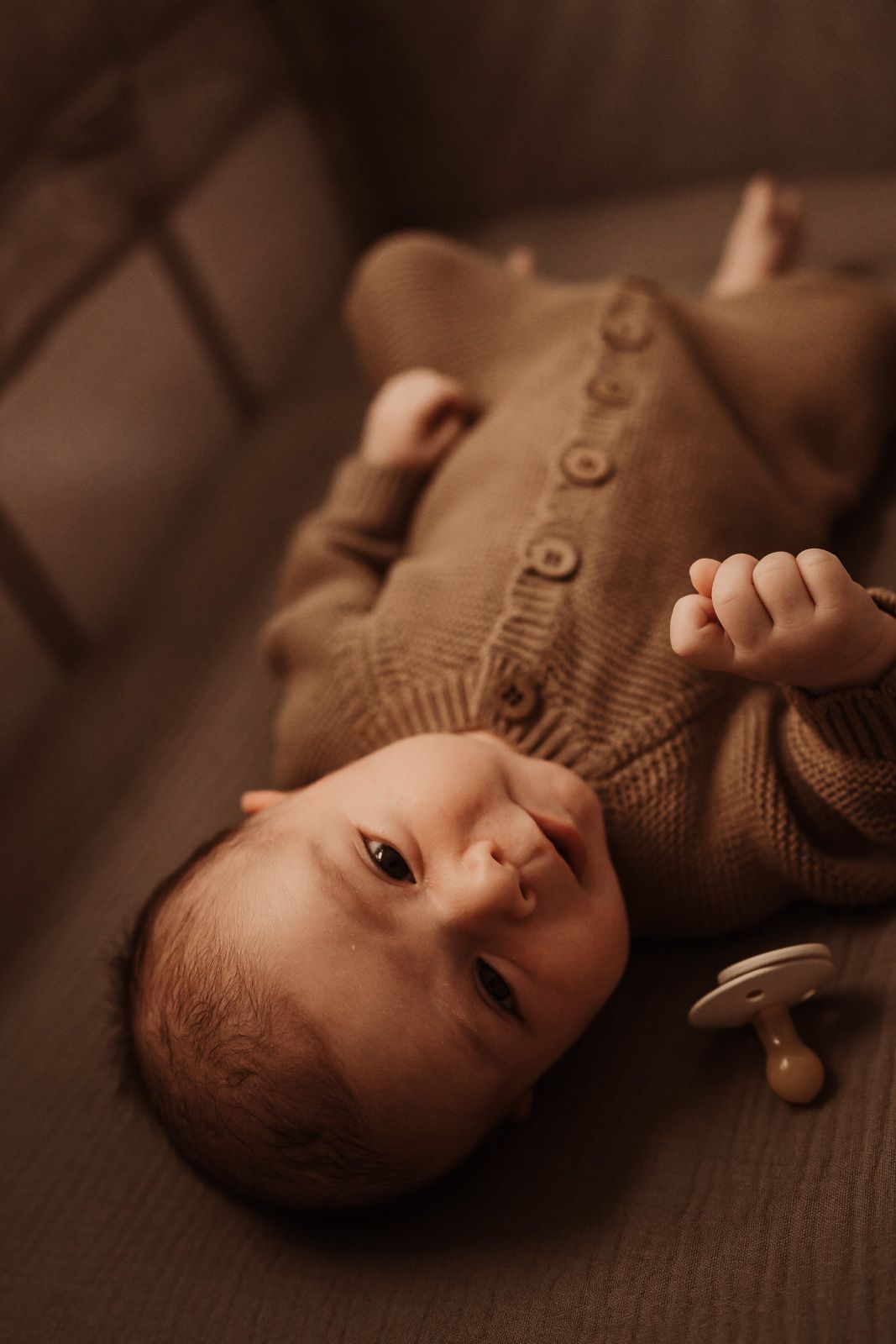 Newborn baby in brown knit onesie lying in crib with pacifier nearby, looking up with dark eyes.