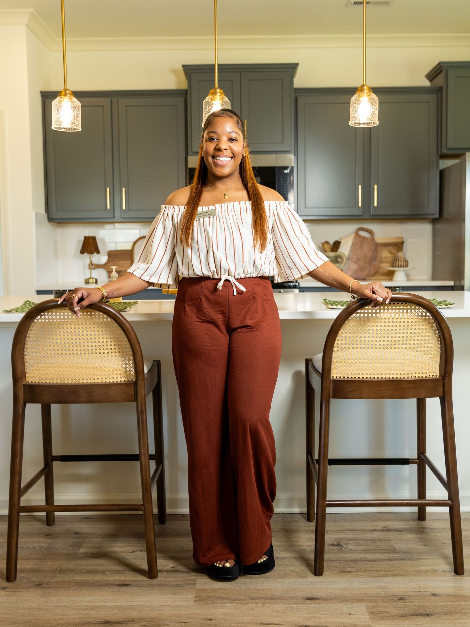 Woman in striped off-shoulder top and rust pants smiling in modern kitchen with dark cabinets and gold accents.