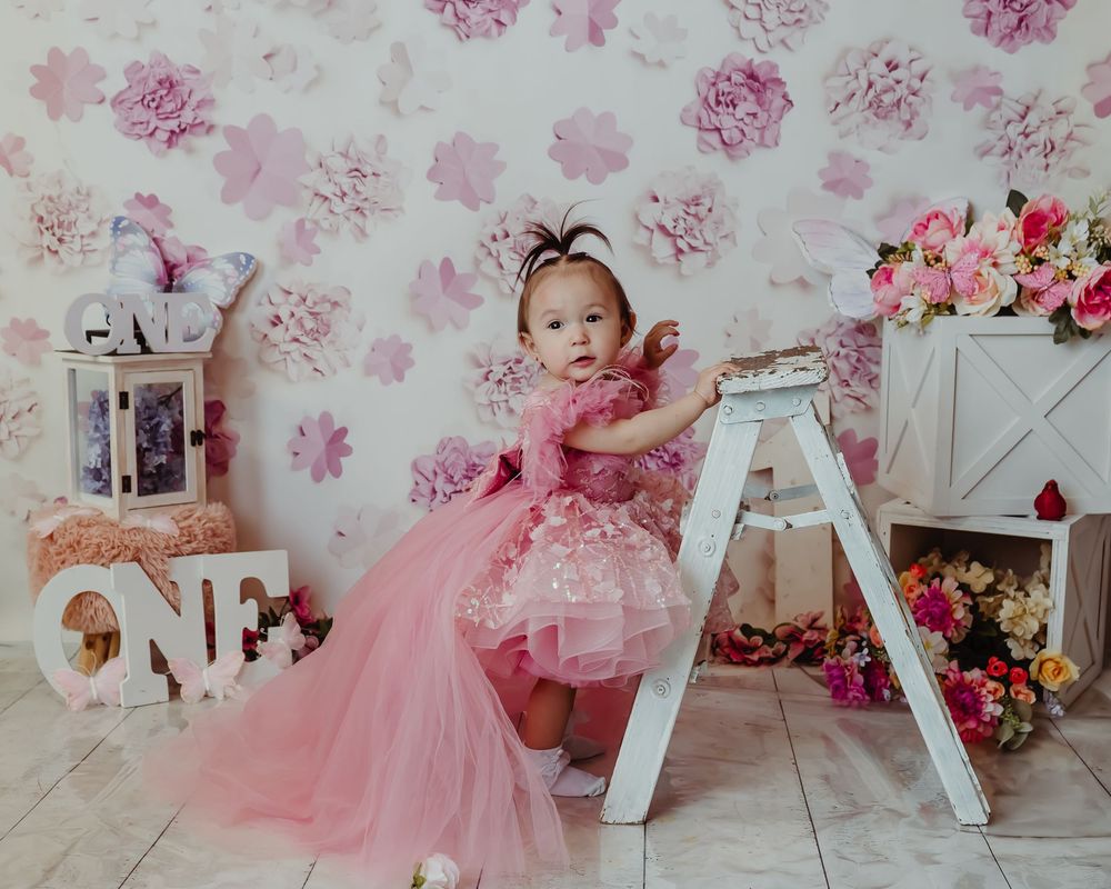 Pink tulle dress against floral backdrop with white ladder, shelves and flower decorations in studio setting.