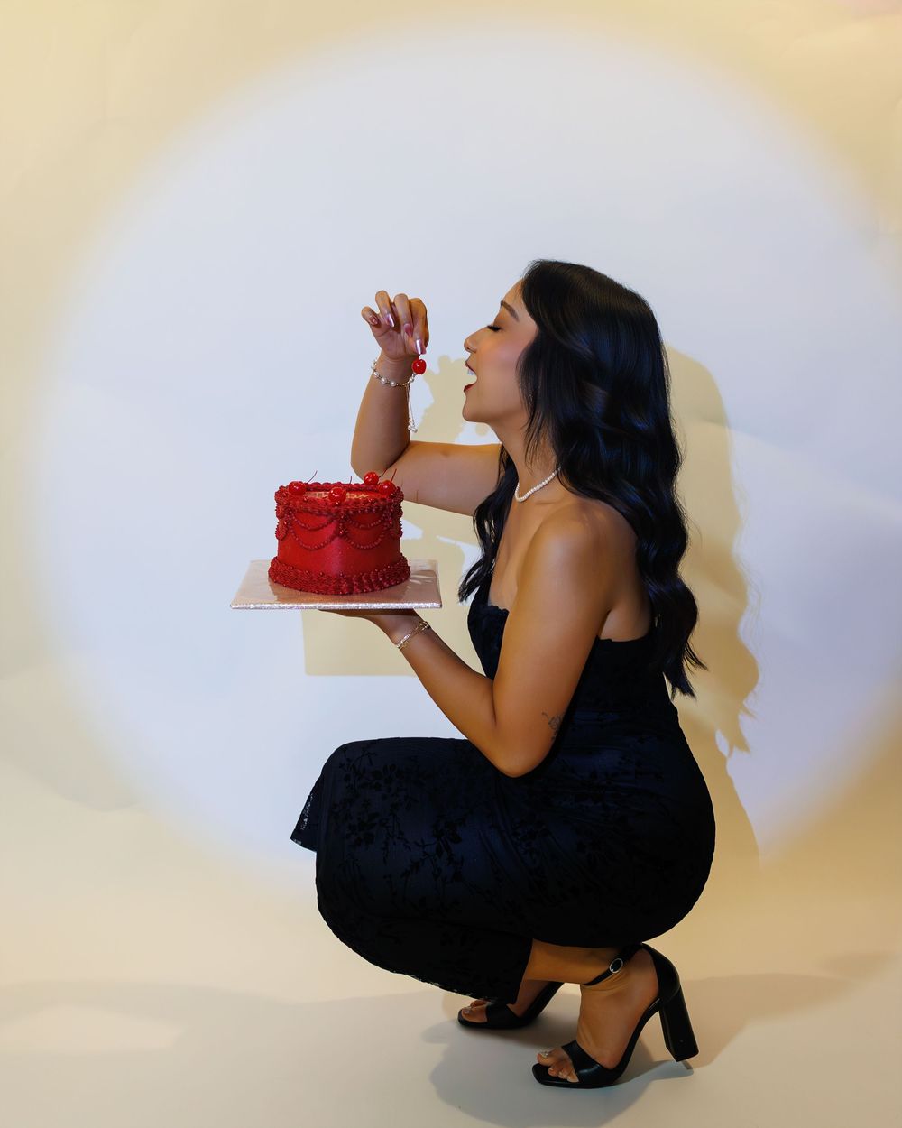 A woman in a black dress poses while holding a bright red cake in front of a white background.