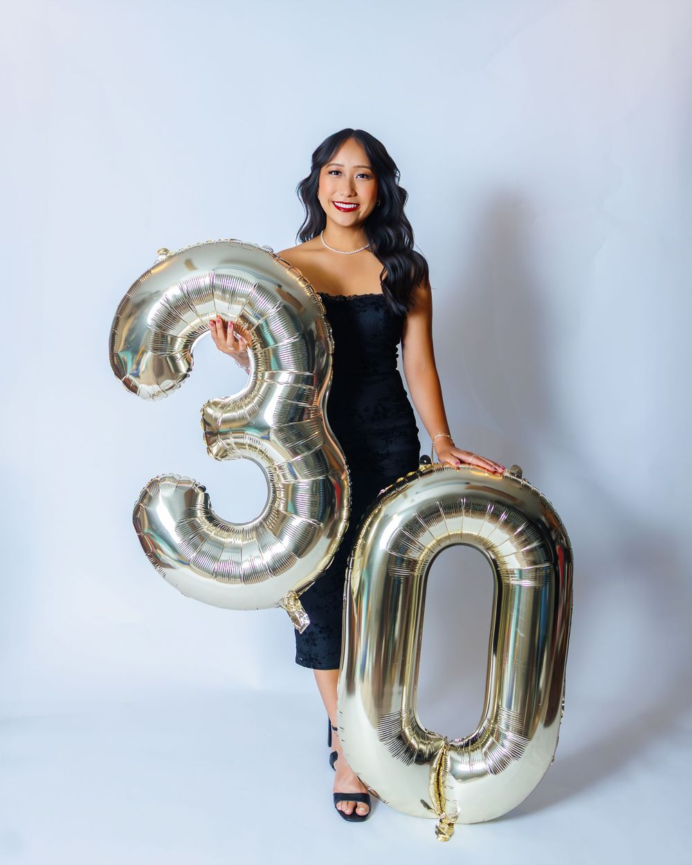 Three photos show a person in a black dress posing with large silver metallic number balloons against a white wall.