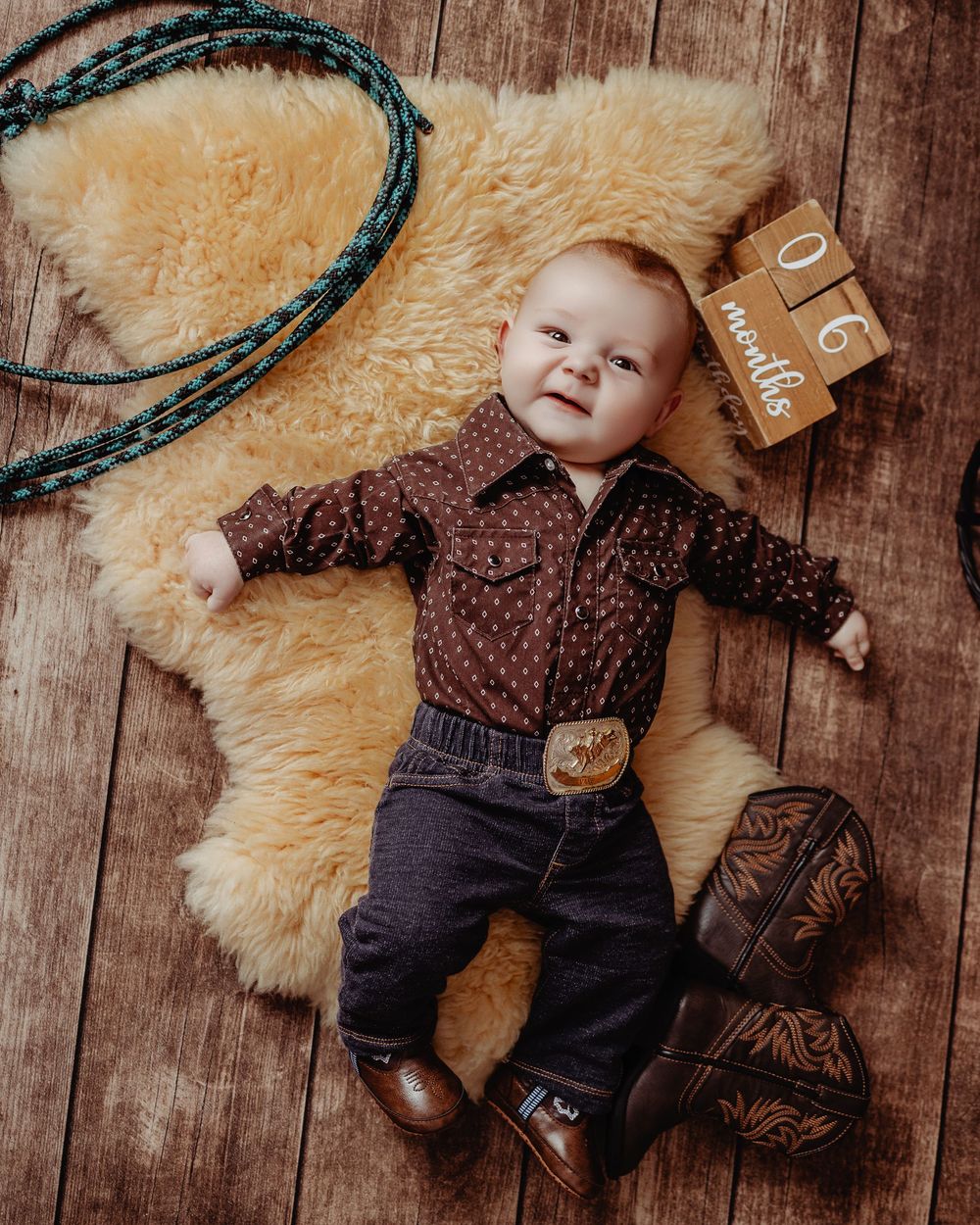 A small child in a western-style outfit lies on a fur rug with cowboy boots and rope on rustic wooden flooring.