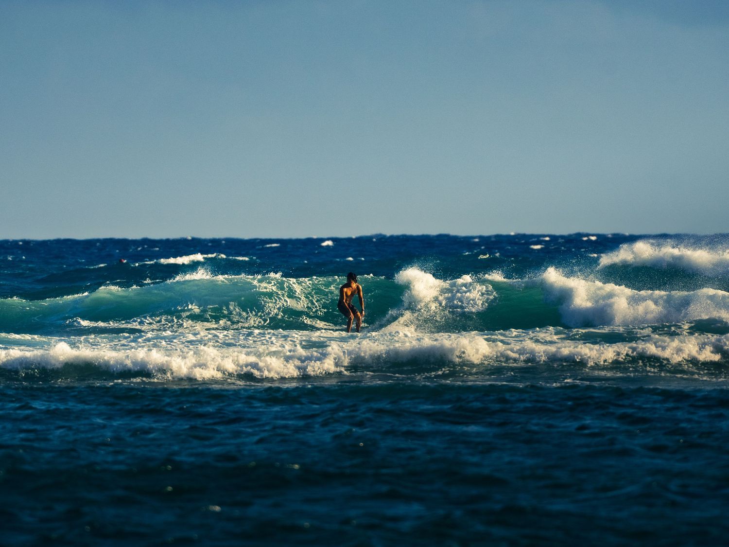 A lone surfer rides a breaking wave across bright turquoise water under a clear blue sky off the coast of Waikiki in Honolulu, Hawaii.