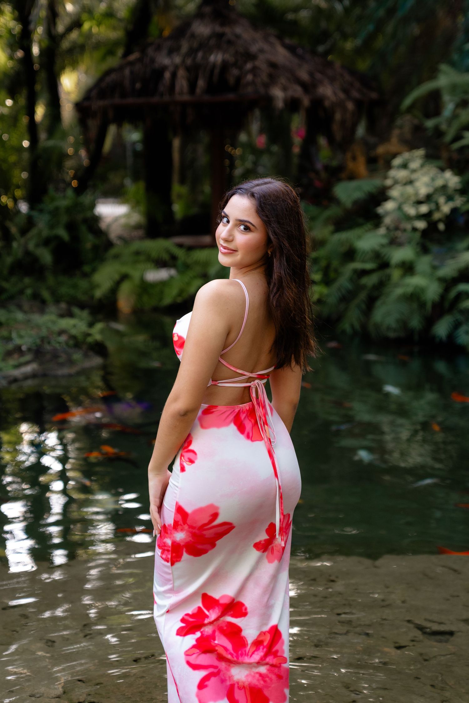Woman in floral backless dress standing in koi pond surrounded by lush tropical garden.