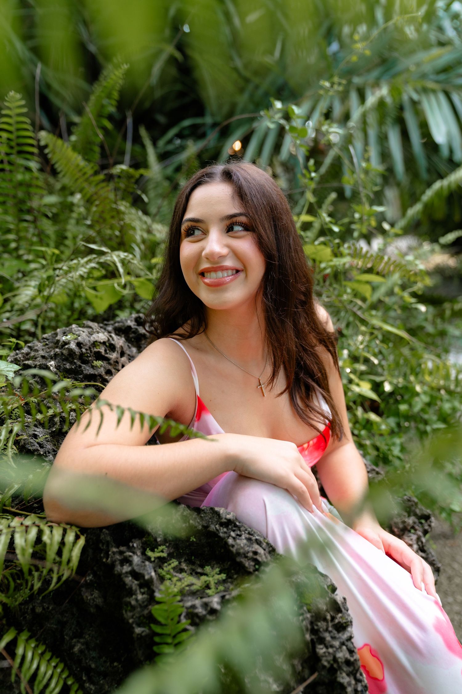 Smiling young woman in a red and pink dress sitting among lush tropical ferns and green foliage.