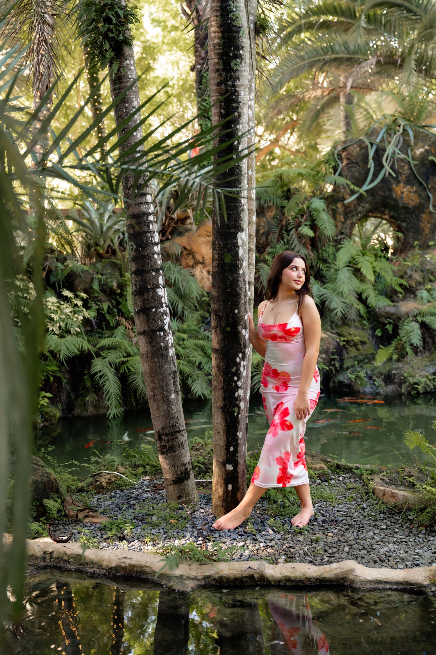 Woman in floral dress standing barefoot by a tranquil pond surrounded by lush tropical palms and ferns.